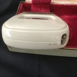 May include: An off-white Remington electric shaver, with the brand name visible on the top. The shaver has a two-pin plug and the voltage is marked as 190-240V. It sits in a green and red velvet lined case.