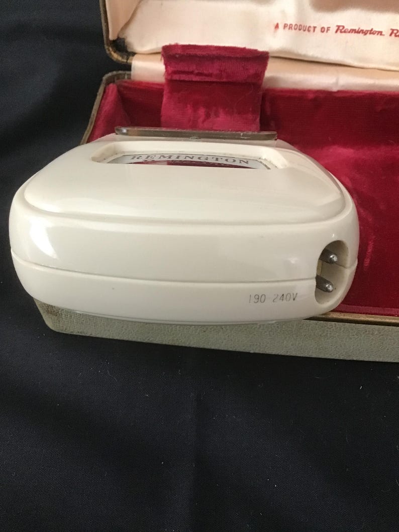 May include: An off-white Remington electric shaver, with the brand name visible on the top. The shaver has a two-pin plug and the voltage is marked as 190-240V. It sits in a green and red velvet lined case.