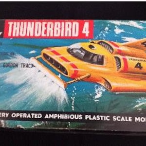 Peut inclure: Une boîte en carton rectangulaire pour un jouet Thunderbird 4. La boîte présente un graphique de véhicule amphibie jaune, le texte A JR 21 TOY, THUNDERBIRD 4, SPECIAL FEATURES, OPERATES AND STEERS IN LAND AND WATER, WITH AQUANAUT GORDON TRACY, et BATTERY OPERATED AMPHIBIOUS PLASTIC SCALE MODEL.