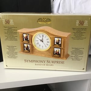 May include: A wooden clock in a box with a gold label that reads "Gold Label Collection". The clock has a round face with Roman numerals and four small framed pictures. The text "Symphony Surprise Band of Bears" is printed on the box.