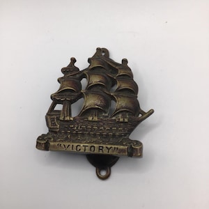 Może przedstawiać: Brązowa kołatka do drzwi w kształcie żaglowca. Kołatka przedstawia szczegółowy statek z żaglami i napisem "VICTORY" na bannerze. Posiada pętlę do zawieszenia.