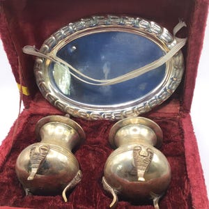 Puede incluir: Un juego de tocador plateado vintage en un estuche forrado de terciopelo rojo. El juego incluye un pequeño espejo ovalado, dos pequeños frascos con diseños de cabeza de elefante y una bandeja plateada. El estuche tiene una cinta amarilla y un cierre.