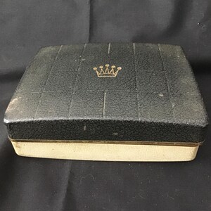 May include: A vintage black jewellery box with a textured lid and a gold crown emblem. The box has a cream-coloured base with a gold trim. The lid is divided into squares.