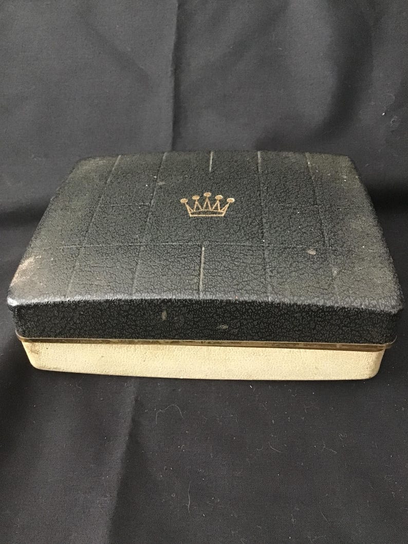 May include: A vintage black jewellery box with a textured lid and a gold crown emblem. The box has a cream-coloured base with a gold trim. The lid is divided into squares.