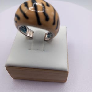 May include: A domed ring with a tan enamel surface and black stripes. The ring is set on a silver band and is displayed on a white and wood-toned jewelry stand. The ring's design resembles a tiger's pattern.