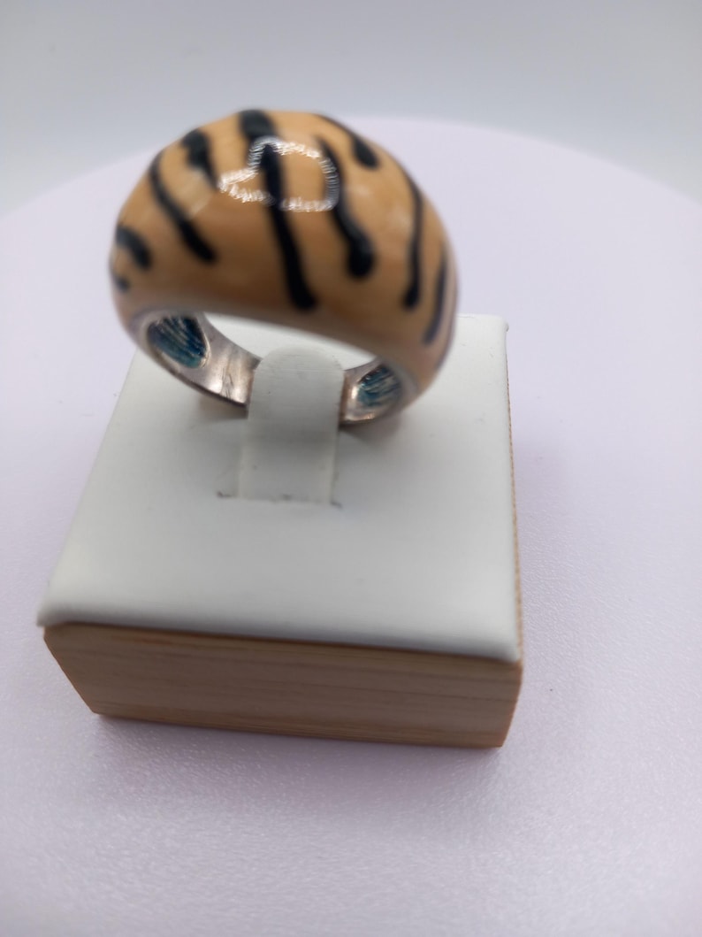 May include: A domed ring with a tan enamel surface and black stripes. The ring is set on a silver band and is displayed on a white and wood-toned jewelry stand. The ring's design resembles a tiger's pattern.