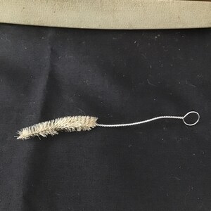 May include: A small cleaning brush with a twisted wire handle and a loop at the end. The brush head is made of light brown bristles. The brush is lying on a black surface.
