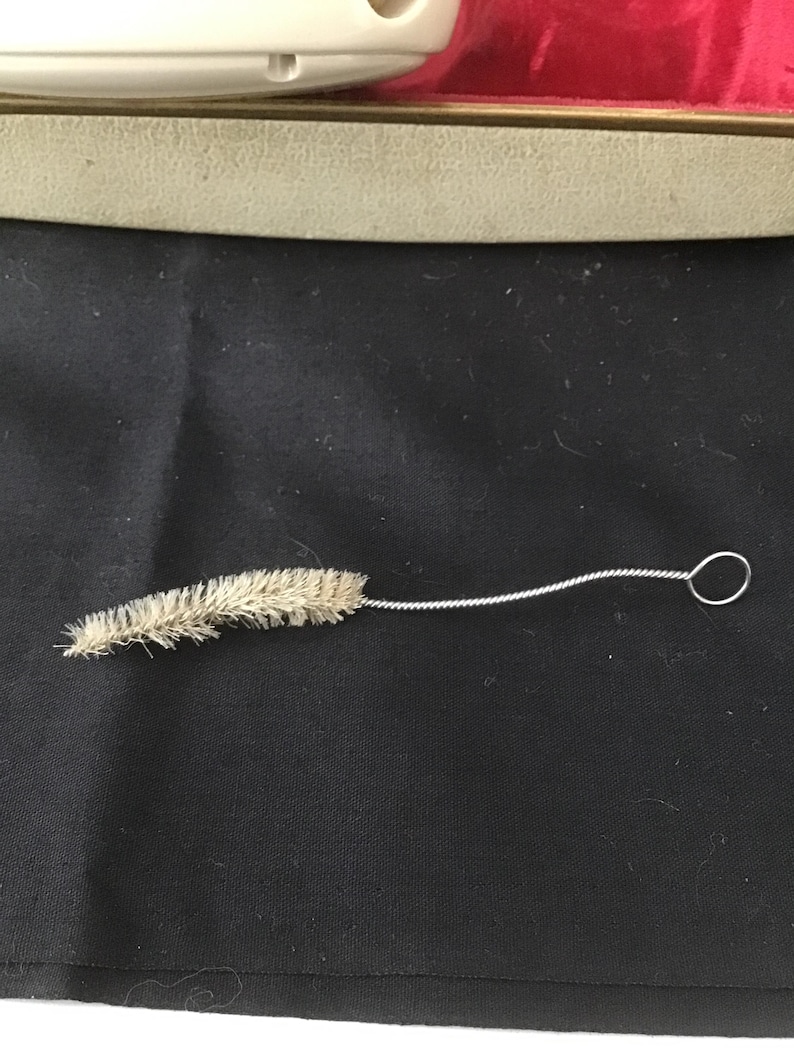 May include: A small cleaning brush with a twisted wire handle and a loop at the end. The brush head is made of light brown bristles. The brush is lying on a black surface.
