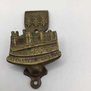 Puede incluir: Un recuerdo de latón vintage del castillo de Caernarvon. La pieza de latón presenta una representación detallada de un castillo, con las palabras "Caernarvon Castle" inscritas debajo. El diseño incluye un escudo en la parte superior y un lazo en la parte inferior.