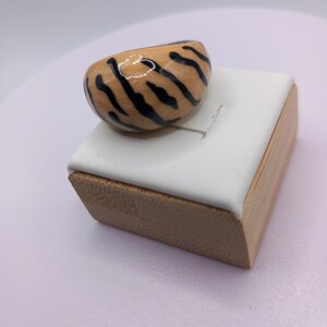 May include: A chunky, tiger-striped ring. The ring features a rounded, oval shape with a light brown base and bold black stripes. It sits on a white and wood-toned display stand, showcasing the ring's unique design and craftsmanship.
