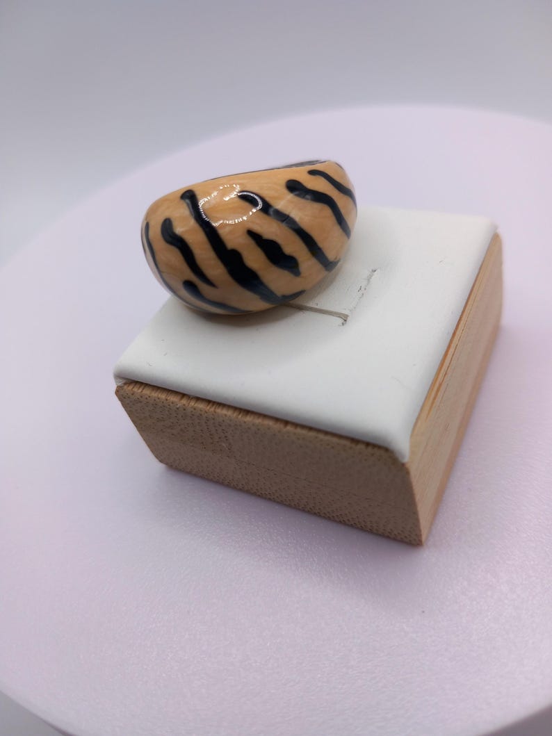 May include: A chunky, tiger-striped ring. The ring features a rounded, oval shape with a light brown base and bold black stripes. It sits on a white and wood-toned display stand, showcasing the ring's unique design and craftsmanship.