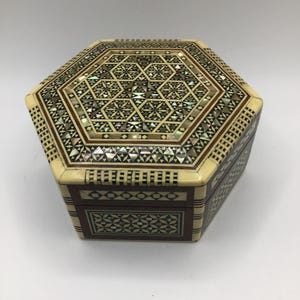 May include: A decorative, hexagonal wooden box with intricate inlaid patterns. The lid features a geometric design with mother-of-pearl accents, while the sides showcase a contrasting dark wood and ivory-colored trim. The box is a beautiful example of craftsmanship.