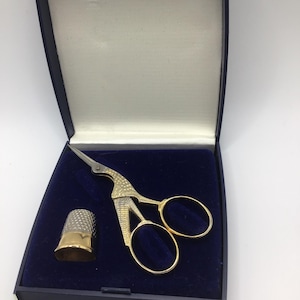 May include: A gold-toned embroidery scissor set with a thimble, presented in a navy blue box. The scissors have a decorative bird-like design. The thimble is gold-colored at the top and silver-colored at the bottom. The box has a white interior.