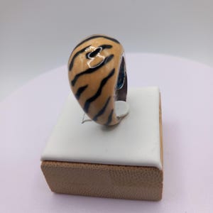 May include: A chunky ring with a tiger stripe design. The ring features a tan base color with black stripes. The ring is displayed on a white and brown jewelry display.