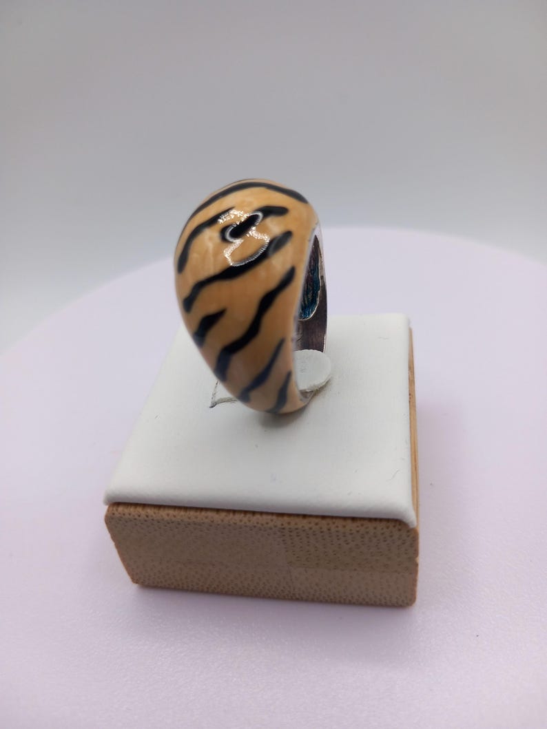 May include: A chunky ring with a tiger stripe design. The ring features a tan base color with black stripes. The ring is displayed on a white and brown jewelry display.