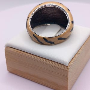 May include: A chunky ring with a cream-colored enamel exterior and black leopard print design. The interior of the ring is dark. The ring is displayed on a white surface atop a wooden box.