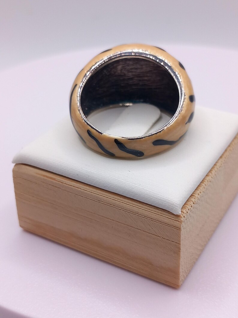 May include: A chunky ring with a cream-colored enamel exterior and black leopard print design. The interior of the ring is dark. The ring is displayed on a white surface atop a wooden box.