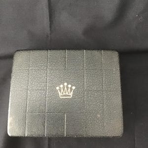 May include: A rectangular, dark grey box with a textured surface and a silver crown emblem in the centre. The box is divided into rectangular sections. The background is a dark grey fabric.