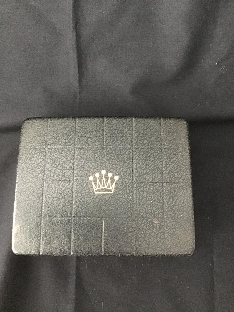 May include: A rectangular, dark grey box with a textured surface and a silver crown emblem in the centre. The box is divided into rectangular sections. The background is a dark grey fabric.