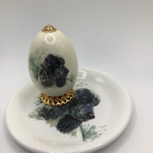 Może przedstawiać: Ozdobna porcelanowa figurka w kształcie jajka z czarno-białym portretem psa. Jajko stoi na pasującym spodku, również ozdobionym portretem psa. Jajko ma złotą filigranową górę i podstawę.