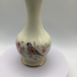 May include: Cream-colored ceramic vase with a gold rim and base. The vase features a painted design of a bird perched on a flowering branch with pink blossoms. The vase is approximately 6 inches tall.