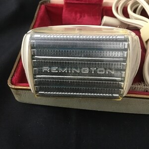 May include: Vintage Remington electric shaver in a case. The shaver has a white and clear plastic housing with the brand name "REMINGTON" visible. The case is lined with red velvet and has a cream-coloured exterior.