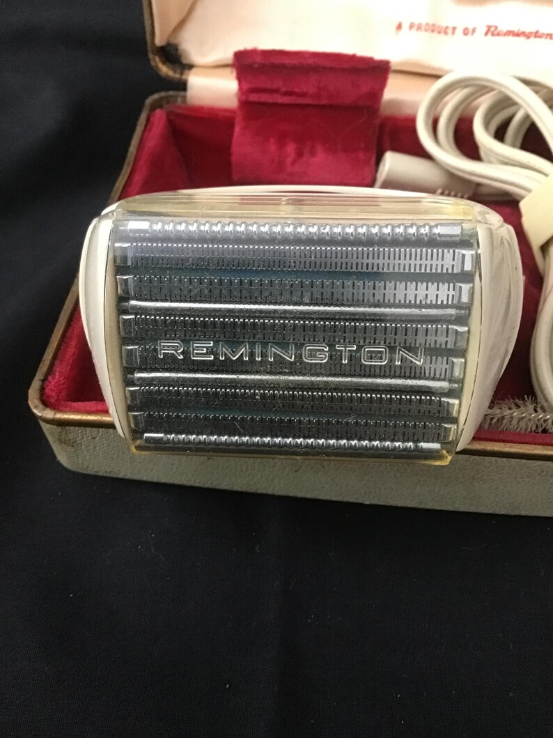 May include: Vintage Remington electric shaver in a case. The shaver has a white and clear plastic housing with the brand name "REMINGTON" visible. The case is lined with red velvet and has a cream-coloured exterior.
