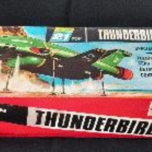 Pode incluir: Uma caixa de brinquedo retangular de papelão para um veículo Thunderbird 2. A caixa apresenta uma ilustração de uma aeronave verde e preta, painéis inferiores vermelhos e texto branco escrito Thunderbird 2 e Friction Motor.