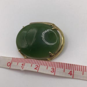 Puede incluir: Una gema redonda y verde engastada en un marco de metal dorado. La gema tiene aproximadamente 2,5 cm de diámetro. Una cinta métrica se coloca cerca de la joya.