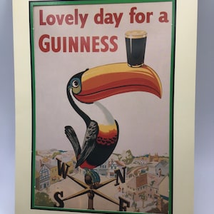 Puede incluir: Impresi&oacute;n enmarcada con un tuc&aacute;n sosteniendo una pinta de Guinness. El texto dice "Lovely day for a Guinness". La imagen incluye una escena de pueblo y una veleta. El marco es verde.