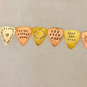 May include: Eight gold and copper guitar picks with various messages engraved on them, including "Rock", "Fly On, Little Wing", "You Rock Dad!" and "Best Dad Ever".