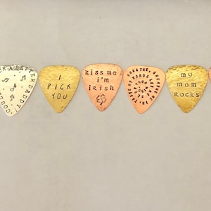 May include: Seven guitar picks with different designs and sayings. The picks are made of metal and have a hammered finish. The sayings on the picks include "I pick you", "Kiss me I'm Irish", "May the music follow you", "My mom rocks", "Breathe the words, believe the feeling", and "You're my heart".