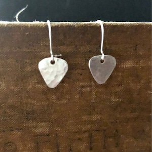 May include: A pair of silver guitar pick earrings with a hammered texture. The earrings are hanging from a brown surface.