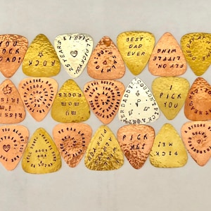 May include: A collection of 18 gold, copper, and silver guitar picks with various engraved messages. The messages include "You Rock Dad!" "Kiss Me, I'm Irish!" "Pick Me!" and more.