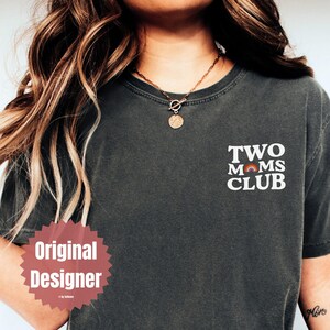 May include: Dark gray t-shirt with the text "TWO MOMS CLUB" in white, featuring a rainbow graphic. The shirt is a casual style, suitable for everyday wear.