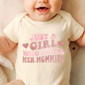 May include: A cream-colored baby onesie with the text "JUST A GIRL WHO LOVES HER MOMMIES" in pink and brown lettering. The onesie has a heart graphic on each side of the word "GIRL".