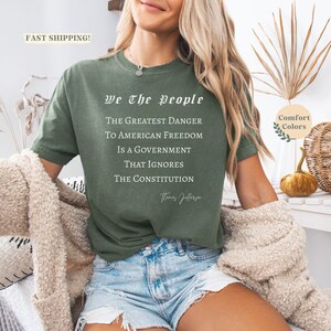 Puede incluir: Una camiseta verde oliva con el texto "We The People" y una cita de Thomas Jefferson. La cita dice: "El mayor peligro para la libertad estadounidense es un gobierno que ignora la Constituci&oacute;n."