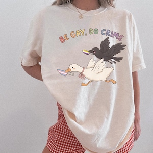 May include: An off-white t-shirt featuring a graphic of a white goose and a black crow, each holding a knife with a colored blade. The text "BE GAY, DO CRIME" is printed in colorful letters above the birds. The goose wears a small pink ribbon.