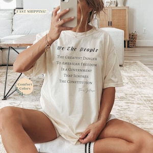 Puede incluir: Camiseta color crema con el texto "We The People" y una cita sobre la libertad estadounidense. La camiseta es de corte holgado y presenta las palabras "Comfort Colors" en la parte inferior izquierda.