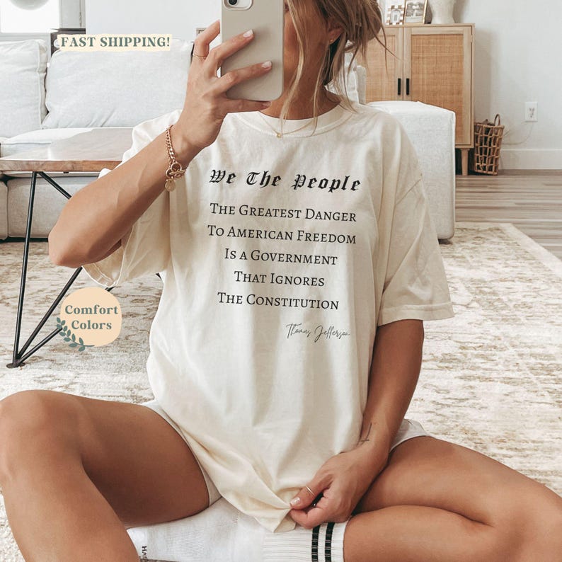 Puede incluir: Camiseta color crema con el texto "We The People" y una cita sobre la libertad estadounidense. La camiseta es de corte holgado y presenta las palabras "Comfort Colors" en la parte inferior izquierda.