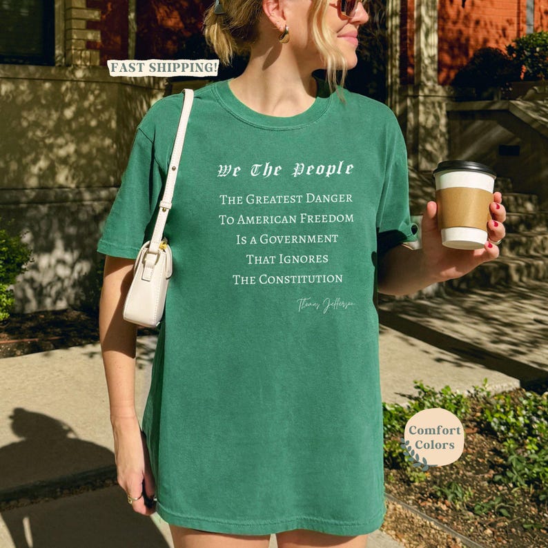 Puede incluir: Una camiseta verde con el texto "We The People" y una cita de Thomas Jefferson sobre la libertad estadounidense. La camiseta es de la marca Comfort Colors.