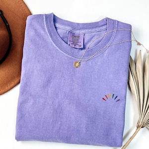 May include: A lavender short-sleeved t-shirt with a ribbed crew neckline and a small embroidered rainbow-colored sunburst design on the lower right chest. The inner neck label reads Comfort Colors, S P CH. A gold-toned necklace with a circular pendant.