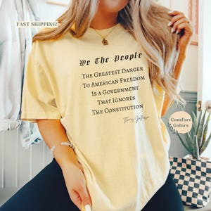 Puede incluir: Una camiseta amarilla clara con el texto "We The People" y una cita de Thomas Jefferson. La camiseta es de la marca Comfort Colors y tiene un corte holgado.