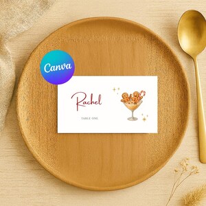 May include: A place card with the name "Rachel" in burgundy script, "Table One" below, and a cocktail illustration with gingerbread men. The card is on a wooden plate with a gold spoon.