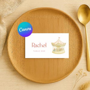 May include: A place setting featuring a wooden plate, gold spoon, and a name card. The name card has the name "Rachel" in red script, the words "Table One", and a carousel illustration. A Canva logo is visible.