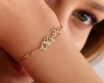 May include: Gold-colored bracelet featuring the name "Chloe" in cursive script, connected to a paw print charm. The bracelet has a delicate chain design. The jewelry is worn on a wrist.