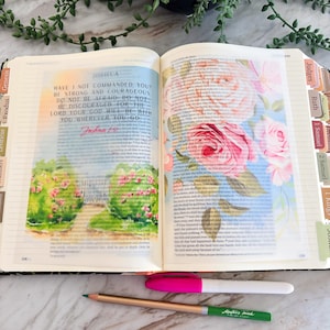 May include: An open Bible featuring a page with the text Joshua 1:9 and a watercolor illustration of a garden path. A translucent vellum overlay displays pink and orange floral patterns. A pink marker and a green pencil lie below the book.