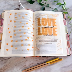 May include: An open Bible with colorful tabs on the edges, featuring a page decorated with orange heart stamps and the text LOVE is patient LOVE is kind 1 Corinthians 13:4 in bold orange and black lettering, accompanied by two yellow pencils.