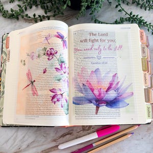 May include: An open Bible with colorful watercolor illustrations of dragonflies and flowers on the pages. The text reads, The Lord will fight for you; You need only to be still. Exodus 14:14. Three pens in pink, purple, and white lie below the book.