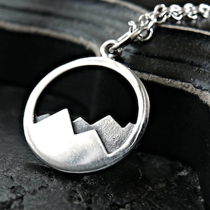 May include: A silver pendant necklace featuring a circular design with a mountain range silhouette. The pendant is attached to a delicate chain. The necklace is displayed on a dark, textured surface.